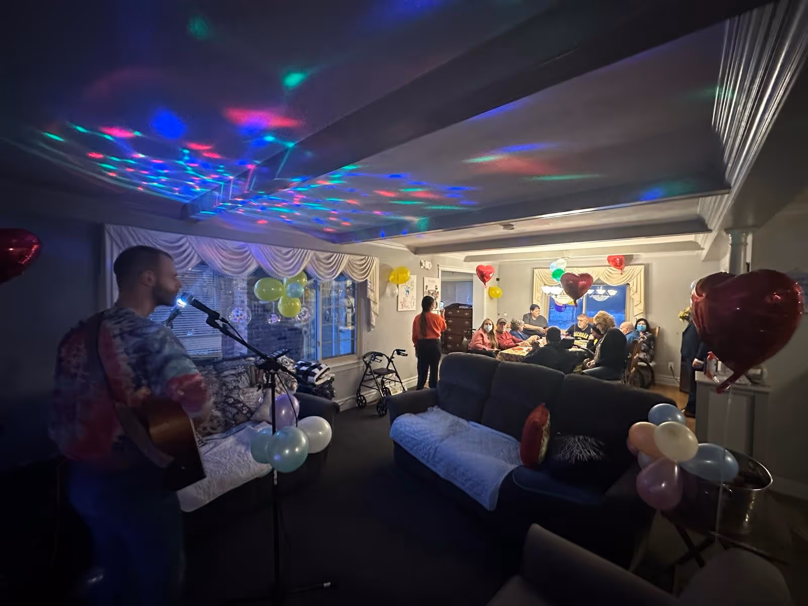 A group of people gathered in a living room decorated with colorful balloons and heart-shaped balloons. A man is playing guitar and singing into a microphone on the left side of the room. The ceiling has colorful disco lights projected on it. Several people are seated on couches and chairs, some wearing masks, enjoying the event.