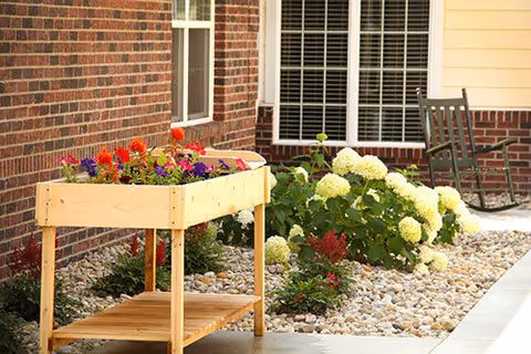 Wooden raised planter filled with colorful flowers beside white hydrangeas and a rocking chair outside a brick building.