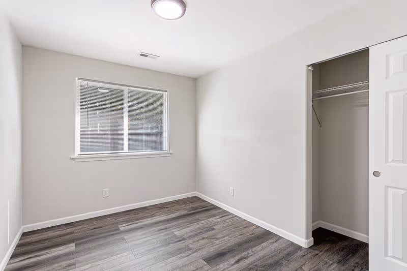 Empty room with light gray walls, a window with blinds, dark wood flooring, a ceiling light, and an open closet with a white door and wire shelving.