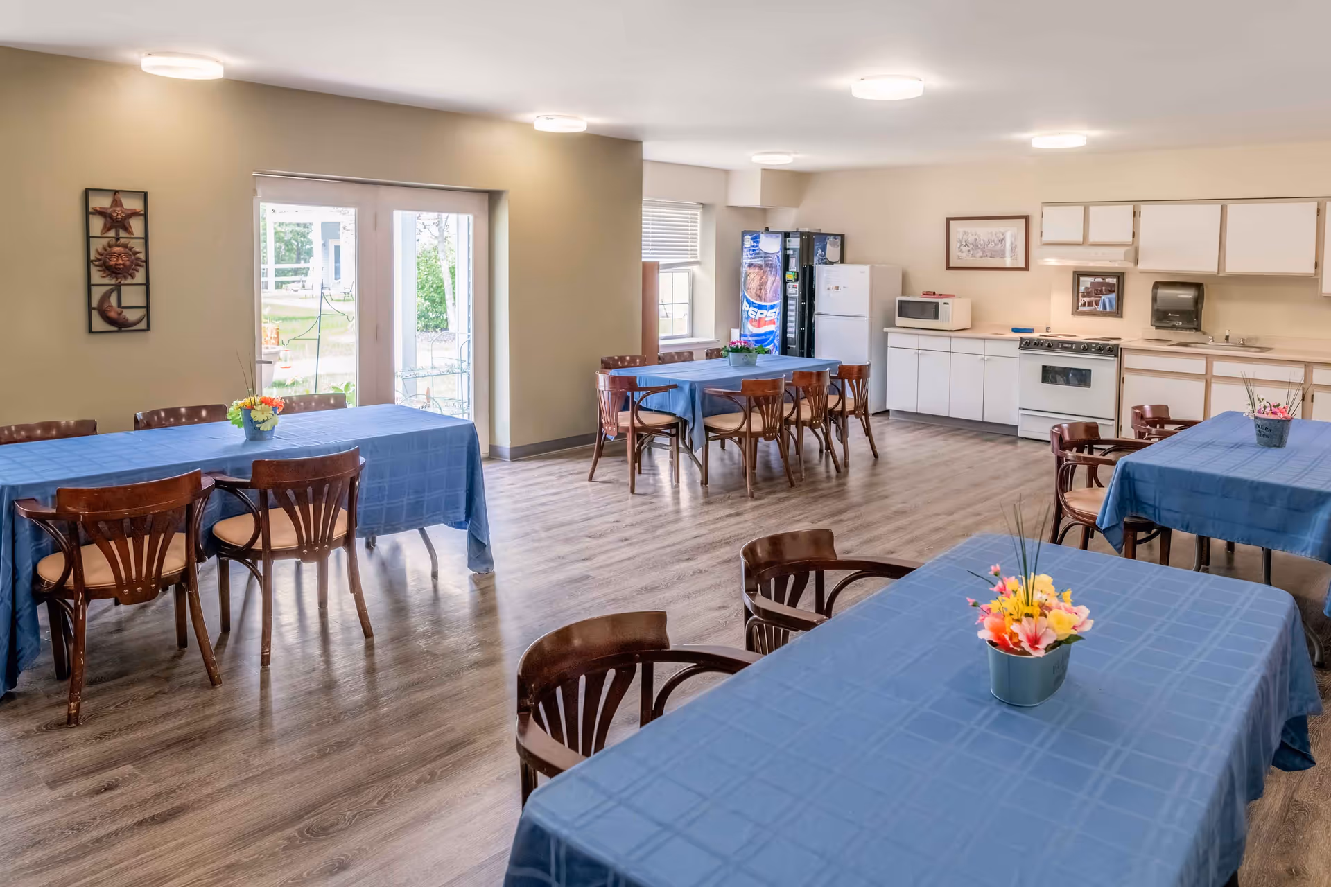 A bright communal dining area with several tables covered in blue tablecloths and decorated with small flower arrangements. Wooden chairs surround the tables. The room features a kitchenette with white cabinets, a stove, microwave, refrigerator, and a Pepsi vending machine. Large windows and glass doors allow natural light to enter, and the floor is covered with wood-patterned vinyl.
