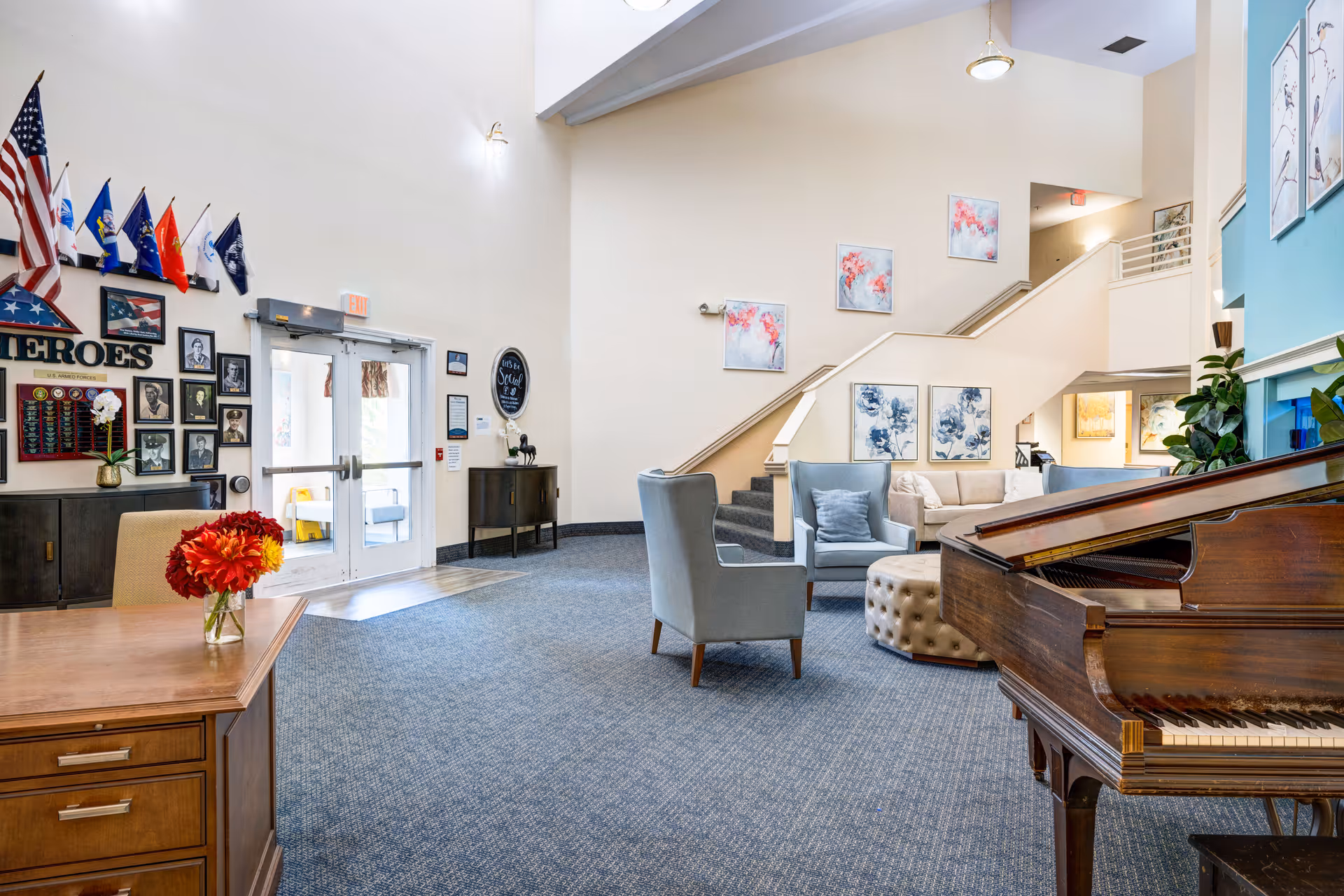 A spacious senior living facility common area with a grand piano on the right, comfortable seating including armchairs and a sofa, a round tufted ottoman, and a staircase leading to an upper level. The walls are decorated with floral artwork and a display of military flags and portraits labeled 'HEROES' near the entrance door. A wooden desk with a vase of red flowers is in the foreground.