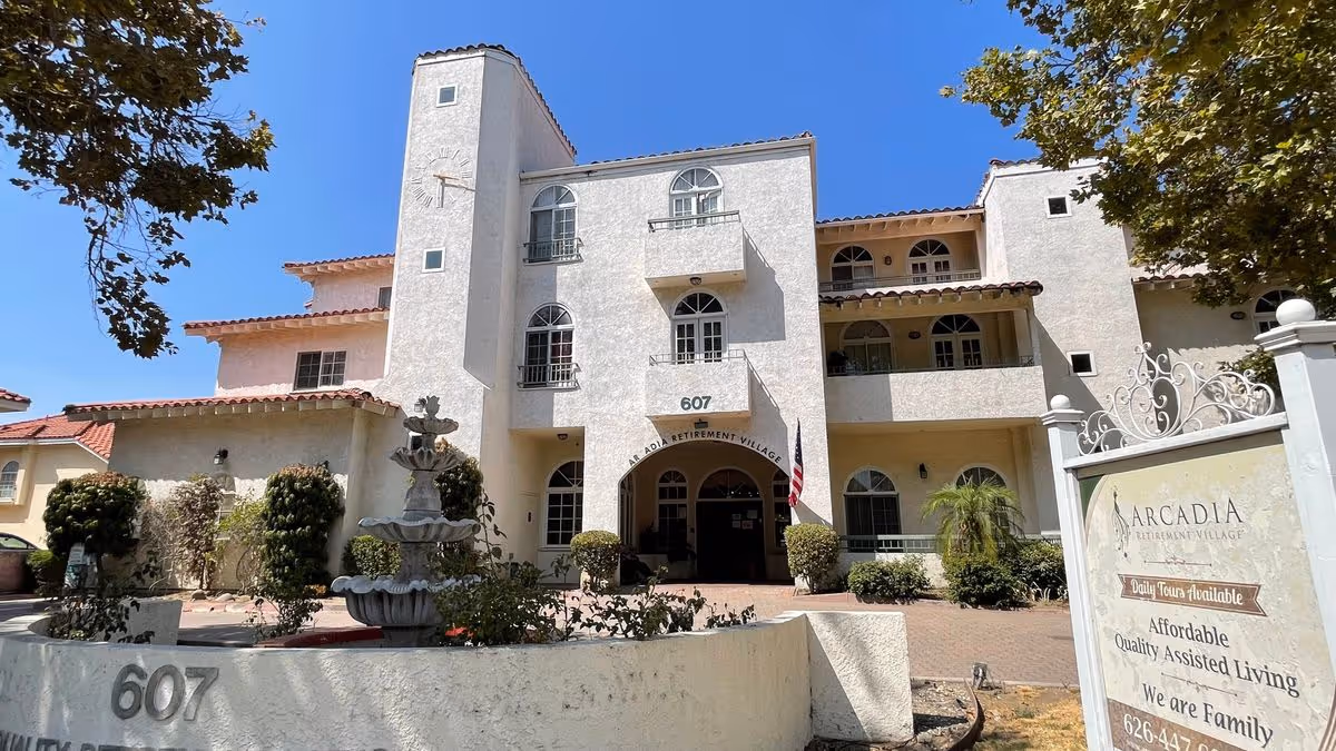 Front entrance of a three-story Mediterranean-style retirement building with a fountain, American flag, and a sign for Arcadia Retirement Village.