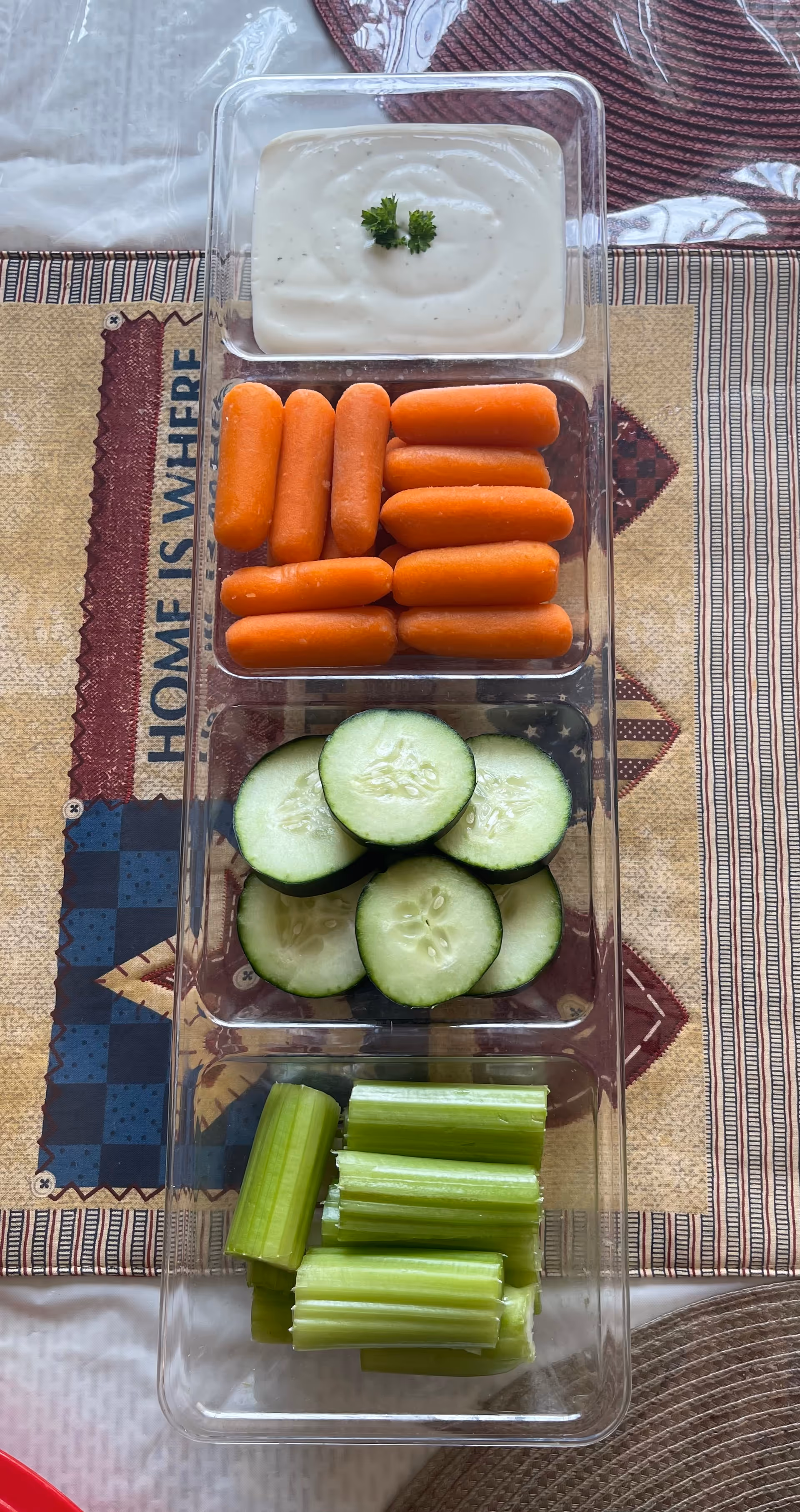 A clear rectangular tray with four compartments containing ranch dressing with a parsley garnish, baby carrots, cucumber slices, and celery sticks, placed on a patterned tablecloth with a design that includes the phrase 'HOME IS WHERE'.