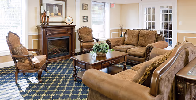 Sunlit communal living room with brown upholstered sofas and armchairs arranged around a wooden coffee table and fireplace.