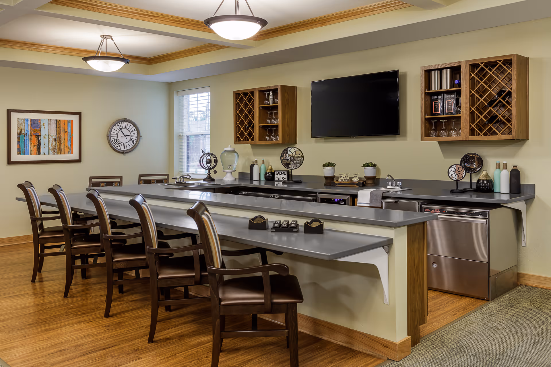 A modern communal kitchen and dining area with a long counter and several wooden chairs with armrests. The counter has a gray surface and is equipped with a dishwasher, sink, and various decorative items including small plants and bottles. Two wooden wall-mounted cabinets with wine racks and glass holders flank a flat-screen TV mounted on a light green wall. The room has wooden flooring, a framed abstract painting, a large wall clock, and two ceiling light fixtures.