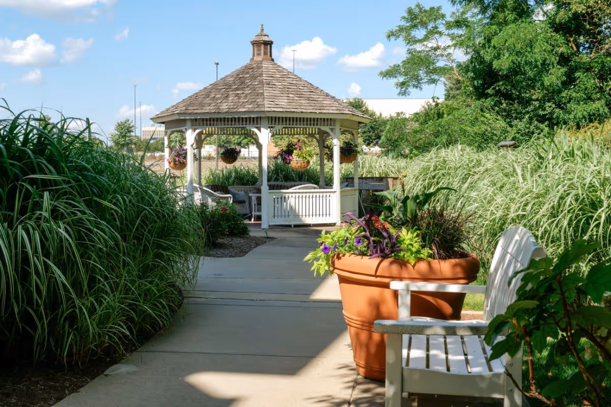 A peaceful outdoor garden area with a paved walkway leading to a white wooden gazebo with a shingled roof. The walkway is flanked by tall green grasses and a large terracotta planter filled with colorful flowers. A white wooden bench is positioned near the planter, and the sky is blue with scattered clouds.