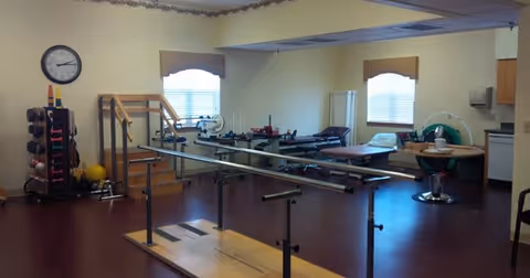 A physical therapy or rehabilitation room with parallel bars for walking exercises, exercise equipment including weights and therapy balls, treatment tables, and a large clock on the wall. The room has windows with blinds and light yellow walls.