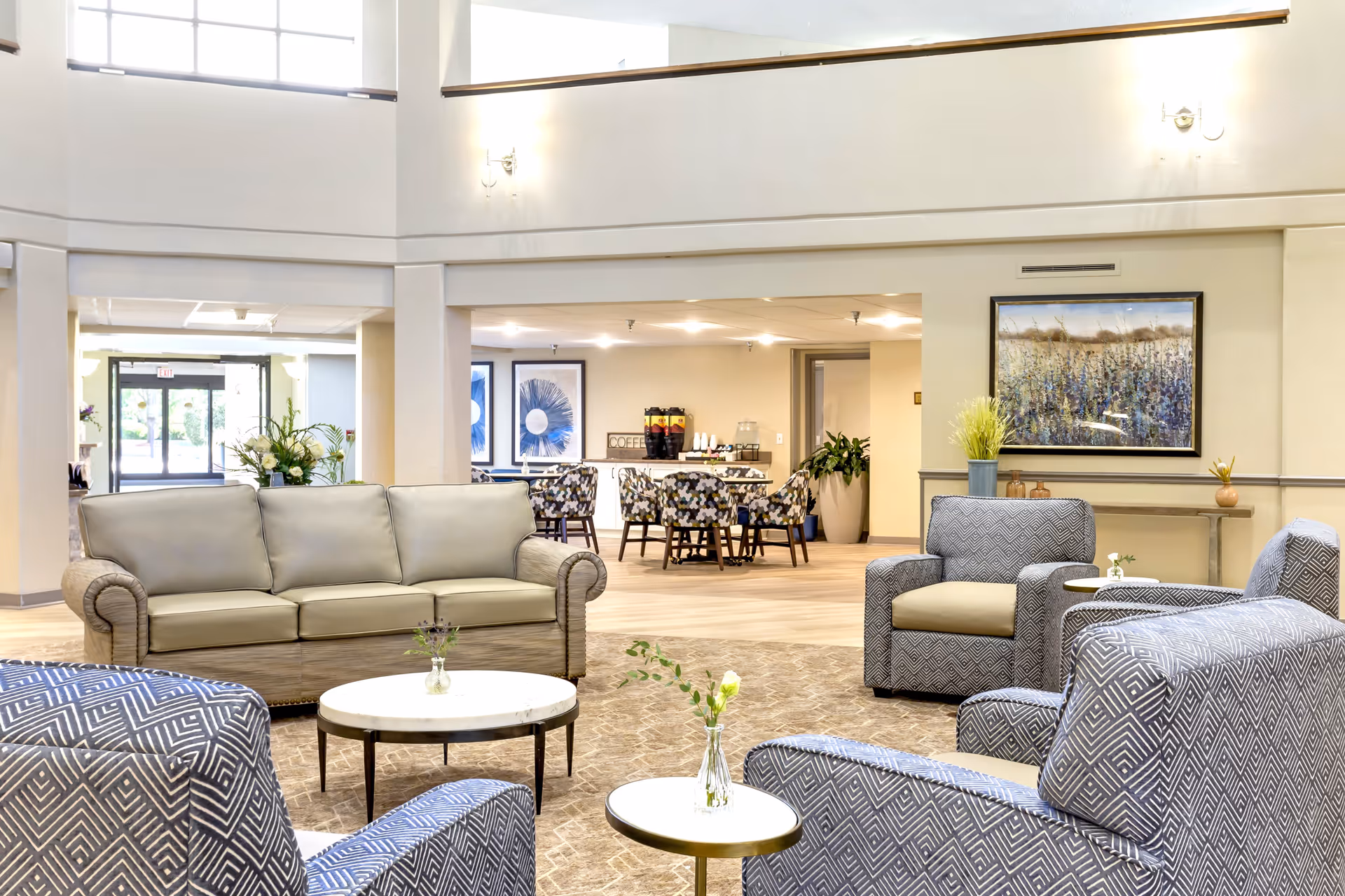A bright and spacious senior living facility common area with comfortable seating including a beige sofa and patterned armchairs arranged around small round tables with flower vases. The background shows a dining area with tables and chairs, coffee dispensers, and decorative artwork on the walls. Large windows and light-colored walls create an airy atmosphere.