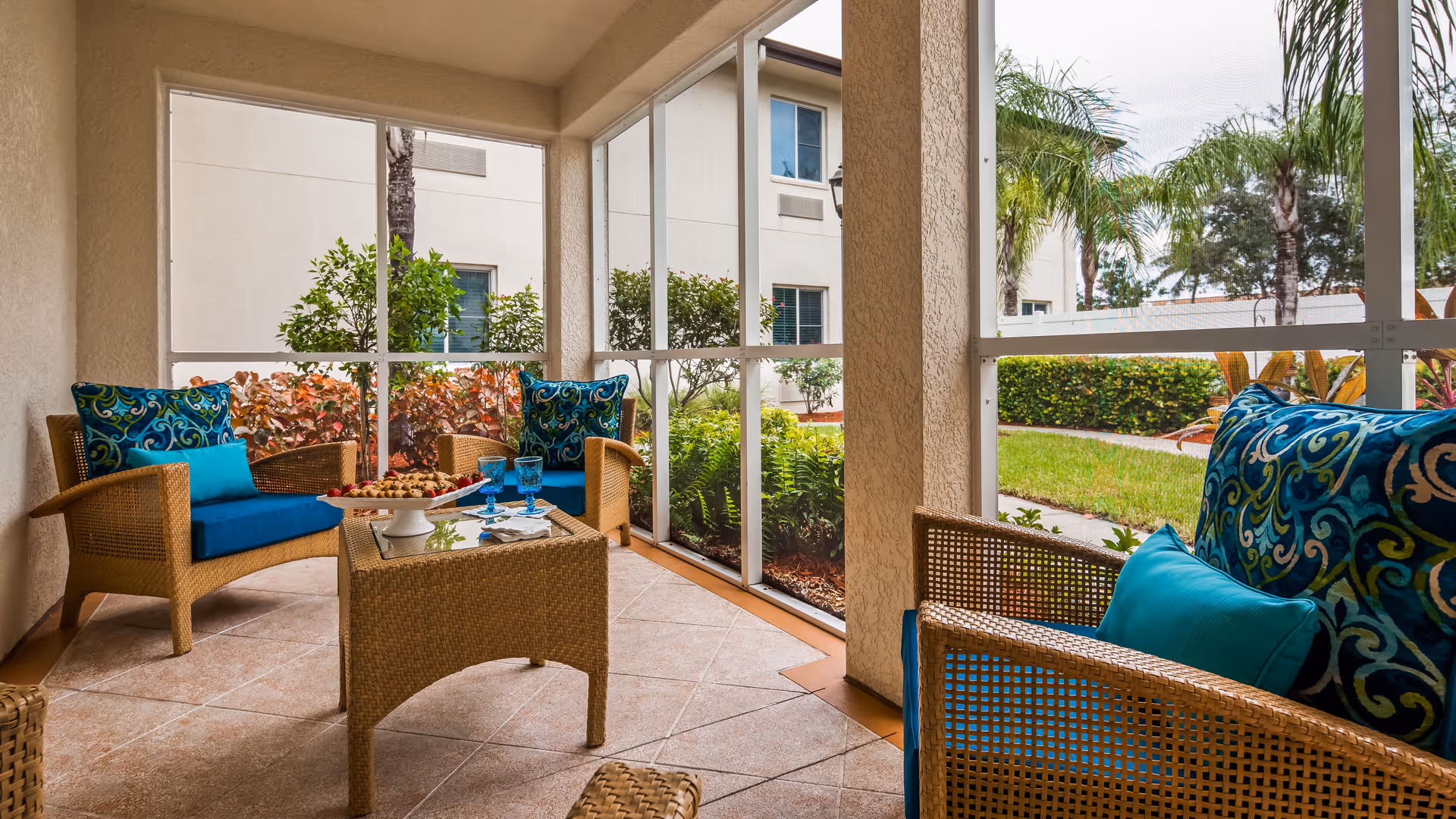 A screened-in patio area with wicker furniture including two armchairs and a loveseat, all with blue cushions and decorative pillows. A wicker coffee table with a glass top holds a plate of cookies and three blue glasses. Outside the screened area, there are green shrubs, palm trees, and a beige building in the background.