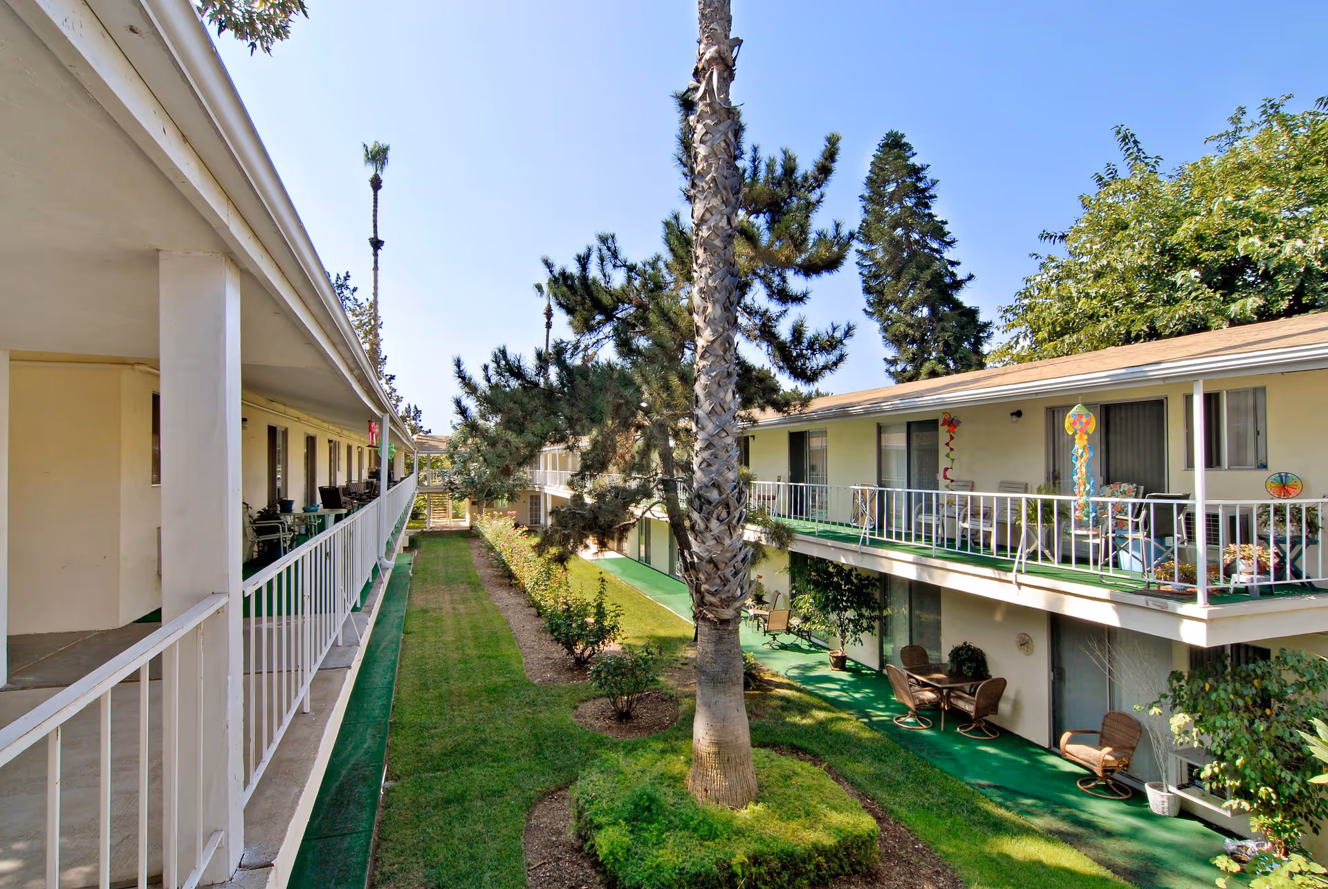 Outdoor view of a senior living facility courtyard with two levels of apartments featuring balconies and patios. The courtyard has green grass, trees including a tall palm tree, and shrubs. The sky is clear and blue.