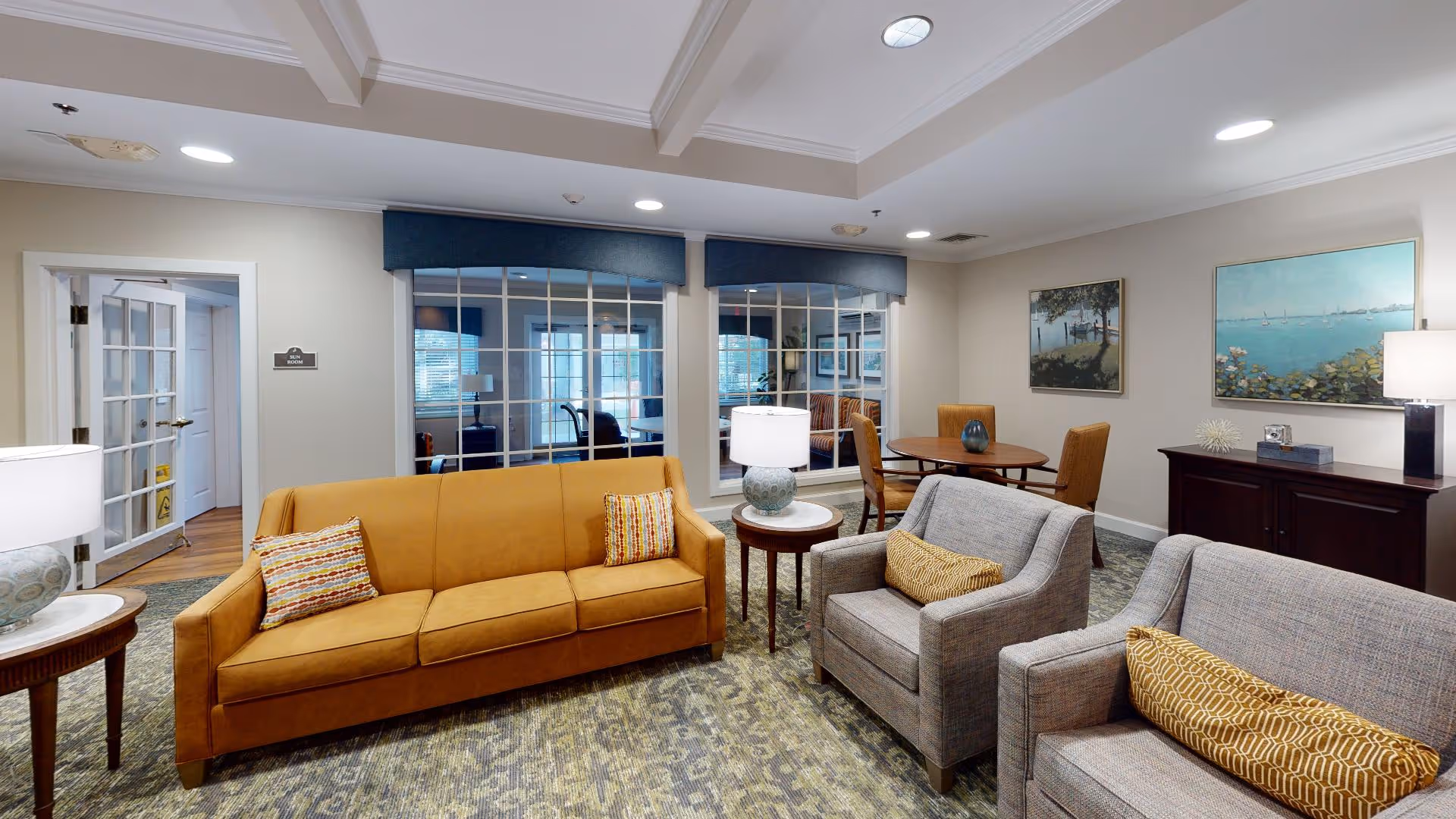 A cozy living room area with a mustard yellow sofa and two gray armchairs, each with decorative pillows. There are two round wooden side tables with lamps on either side of the sofa. In the background, a round wooden table with four chairs is visible, along with framed artwork on the walls and large windows with blue valances.