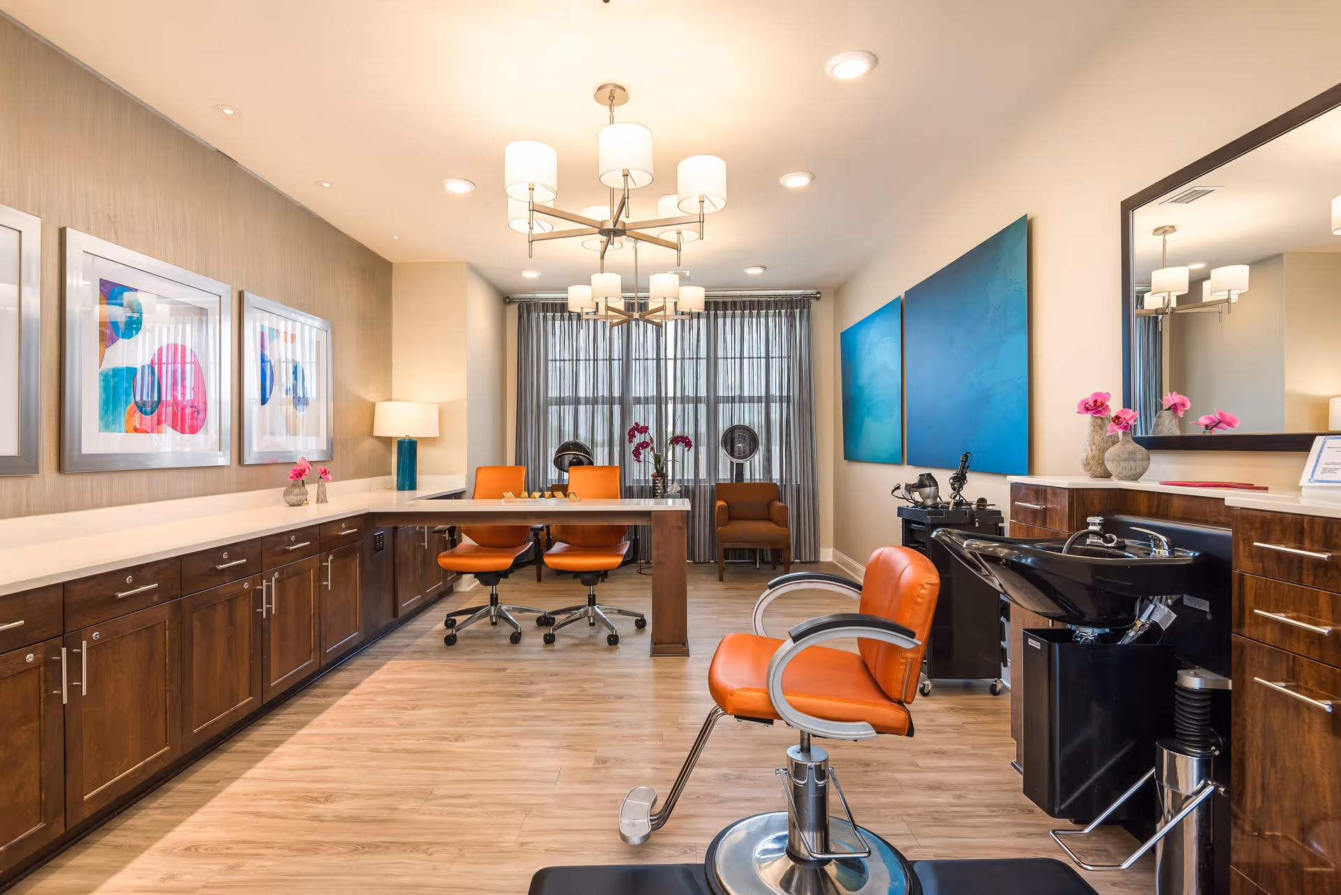 A bright and modern salon area in a senior living facility with orange salon chairs, a hair washing station, wooden cabinets, colorful abstract artwork on the walls, a large mirror, and a window with sheer curtains letting in natural light.