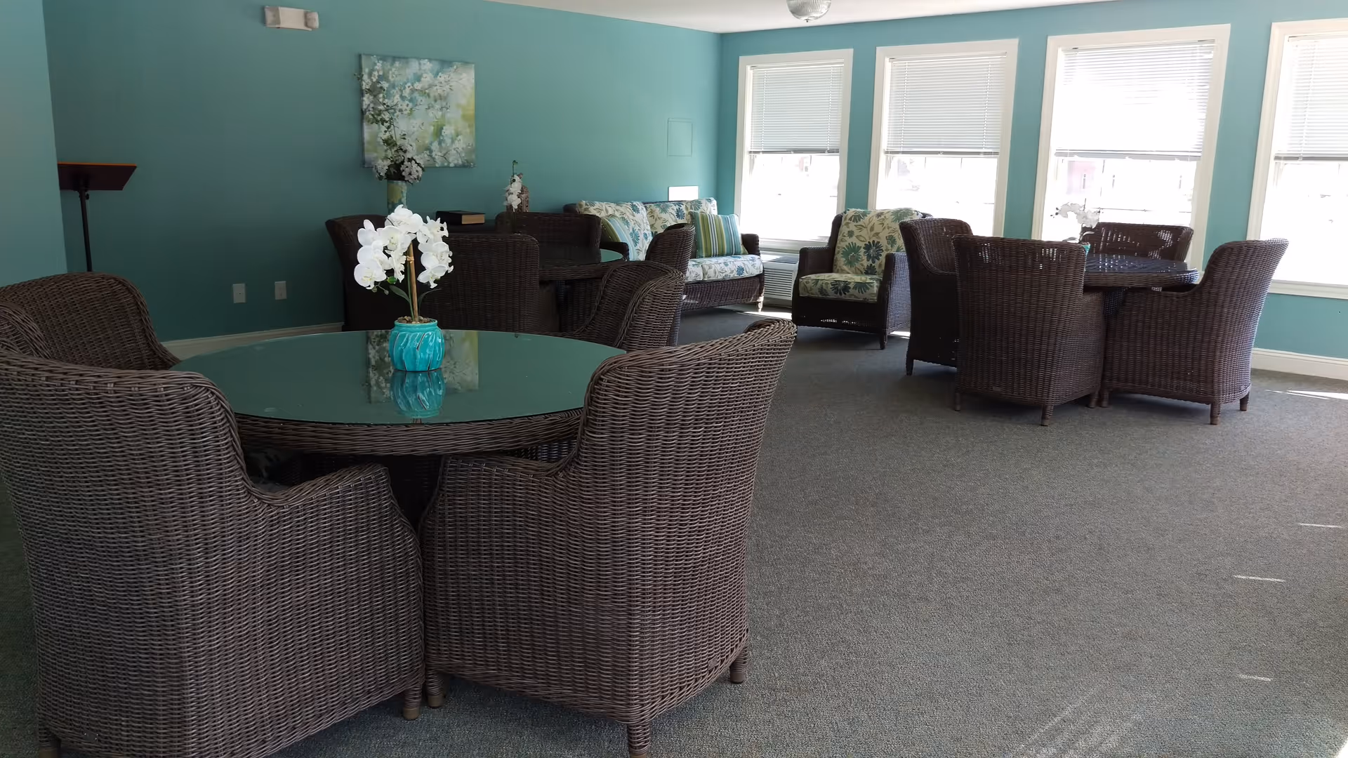 A bright common area with teal walls and multiple wicker seating arrangements including round tables with chairs and cushioned armchairs. There are large windows letting in natural light, and a small vase with white flowers is placed on the glass tabletop in the foreground.