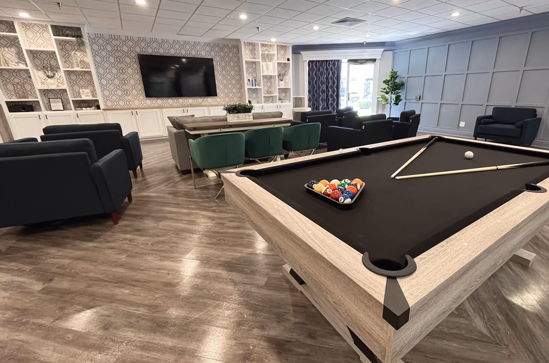 A spacious common area with a pool table in the foreground, two pool cues and a rack of billiard balls on the table. The room features multiple seating options including dark blue armchairs, green chairs, and a sofa facing a large flat-screen TV mounted on a patterned wall with built-in shelves and cabinets. The floor is wood-style, and there is a large window with dark curtains letting in natural light.