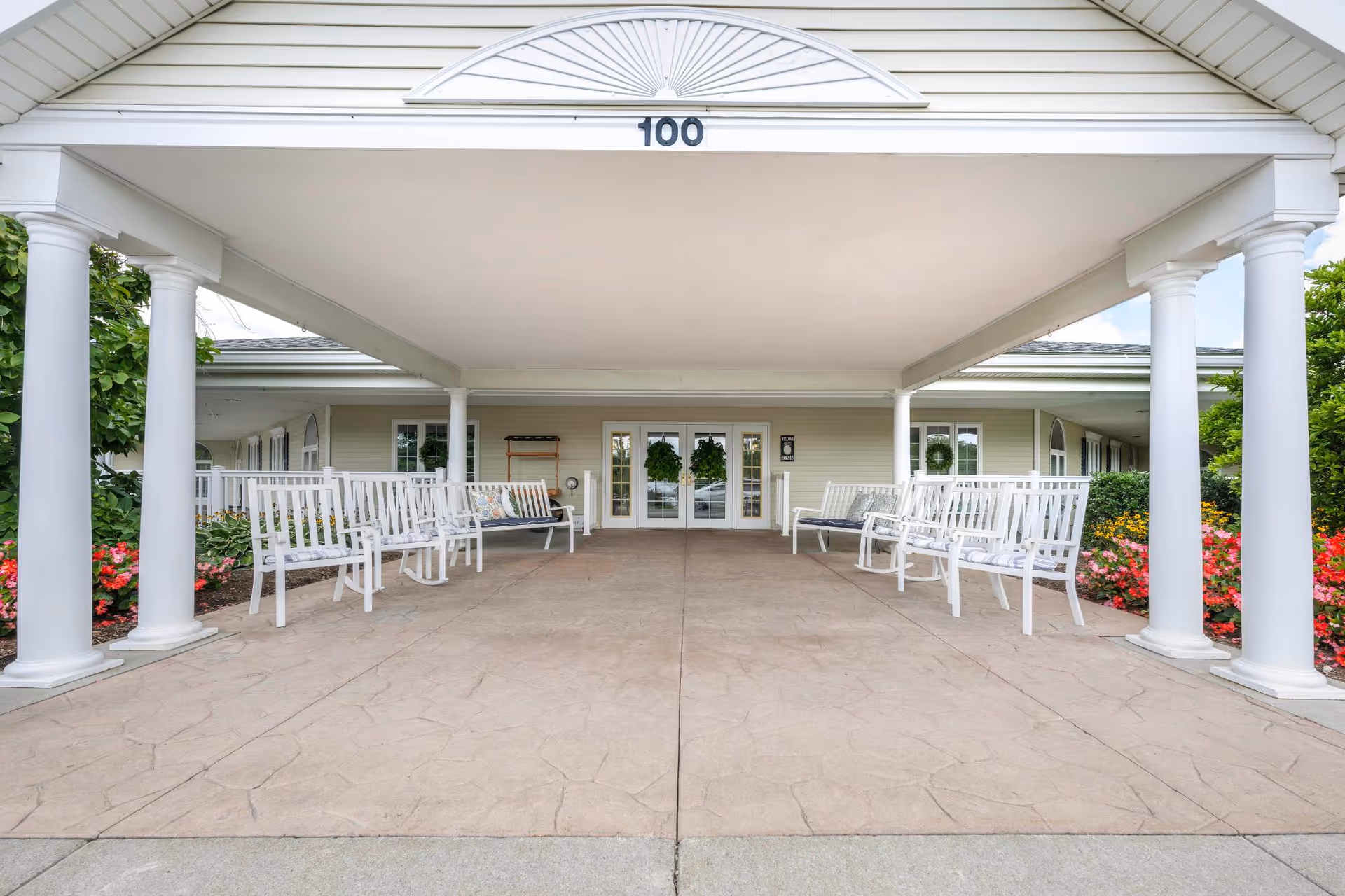 Covered entrance area of a senior living facility with white columns and white benches on both sides. The building has double glass doors decorated with green wreaths, and there are colorful flower beds on either side of the entrance.