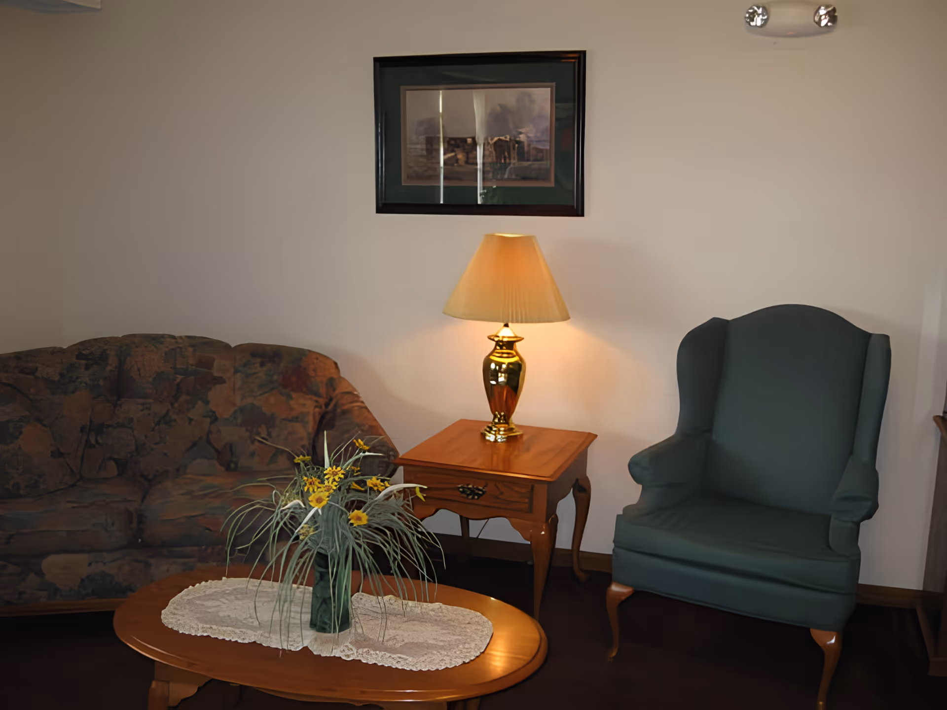 A cozy living room area with a patterned sofa on the left, a green upholstered wingback chair on the right, and a wooden coffee table with a lace doily and a vase of yellow flowers in the center. A wooden side table with a gold lamp and a beige lampshade is positioned between the sofa and the chair. A framed picture hangs on the wall above the side table.