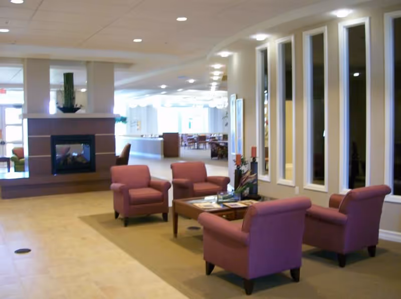 A cozy seating area in a senior living facility with four pink upholstered armchairs arranged around a wooden coffee table. The space features a fireplace with a plant on top, large vertical windows on the right wall, and a bright dining area visible in the background.