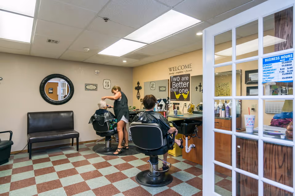 Interior of a small salon area in an assisted living facility with a hairstylist cutting an elderly woman's hair. Another person is seated in a salon chair facing a mirror. The room has checkered flooring, a bench against the wall, a round mirror, and a sign on the wall that reads 'Two are Better than one'. A glass door with business hours posted is visible on the right side.
