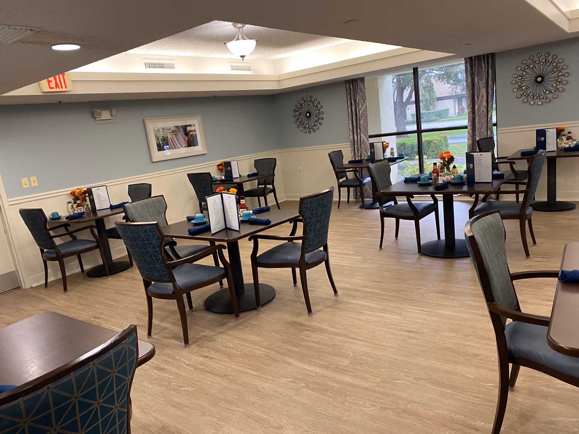 A dining room with several dark wood tables and chairs arranged neatly. Each table has menus, blue napkins, blue cups, and small flower arrangements. Large windows with patterned curtains allow natural light to enter, and there are decorative wall hangings and a framed picture on the light blue walls.