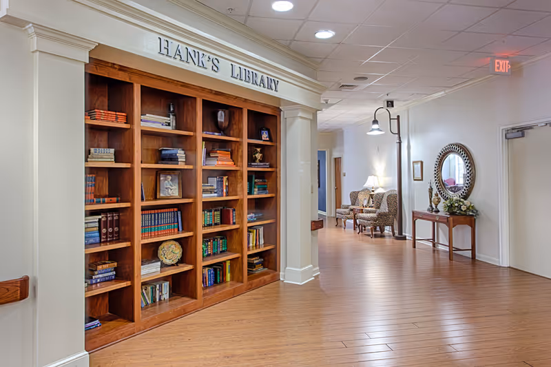 Interior hallway of an assisted living facility featuring a wooden bookshelf labeled 'Hank's Library' filled with books and decorative items. The hallway has wooden flooring, a small table with a flower arrangement and a round mirror on the wall, two patterned armchairs with a lamp in the background, and an exit sign above a door.
