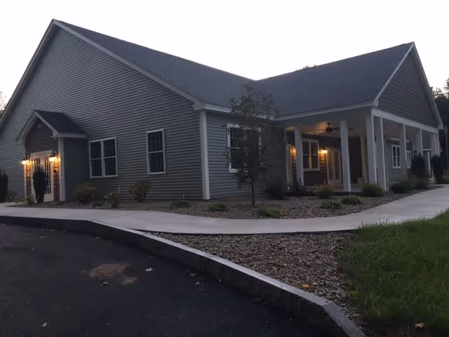 Front exterior of a one-story gray building with a covered porch, lit entrance, and a curved concrete walkway.