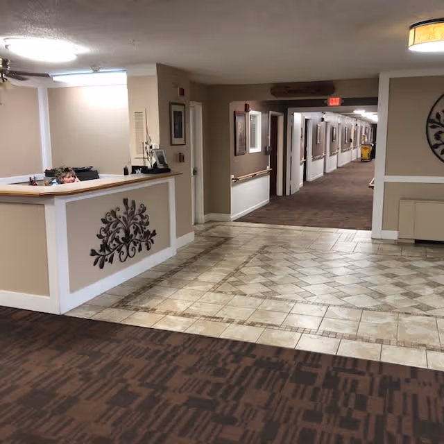 Reception area with a front desk on the left side, decorated with a metal wall art piece. The floor transitions from patterned carpet to tiled flooring leading to a hallway with handrails and framed pictures on the walls. A cleaning cart is visible in the hallway under an exit sign.
