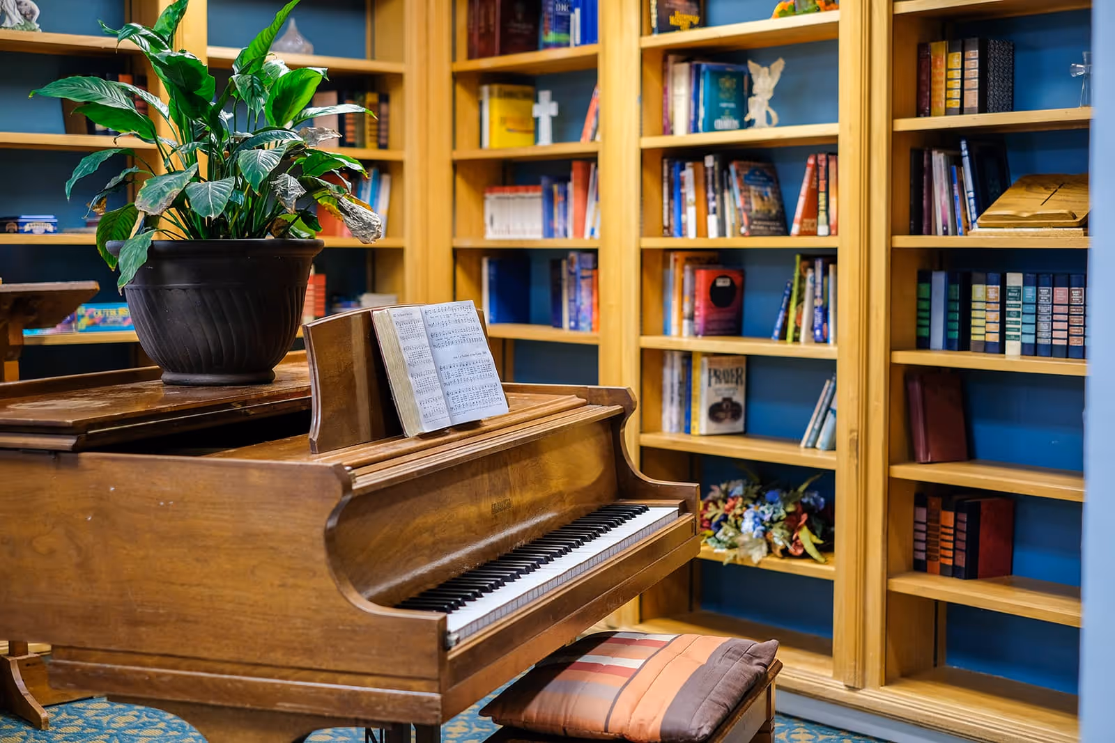 A wooden piano with sheet music on the stand and a potted plant on top, situated in front of wooden bookshelves filled with books and decorative items in a cozy room.