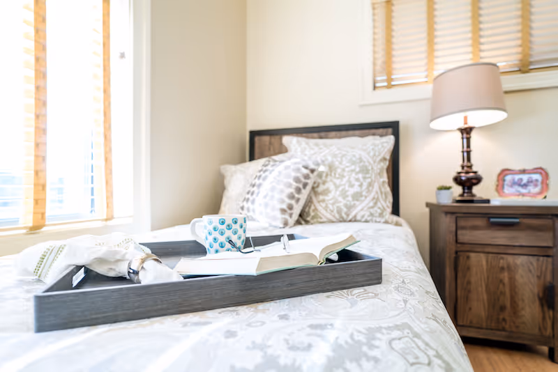 A neatly made bed with patterned pillows and a tray holding a coffee mug, an open book, and a napkin. Next to the bed is a wooden nightstand with a lamp, a small plant, and a framed photo. Sunlight streams in through window blinds.
