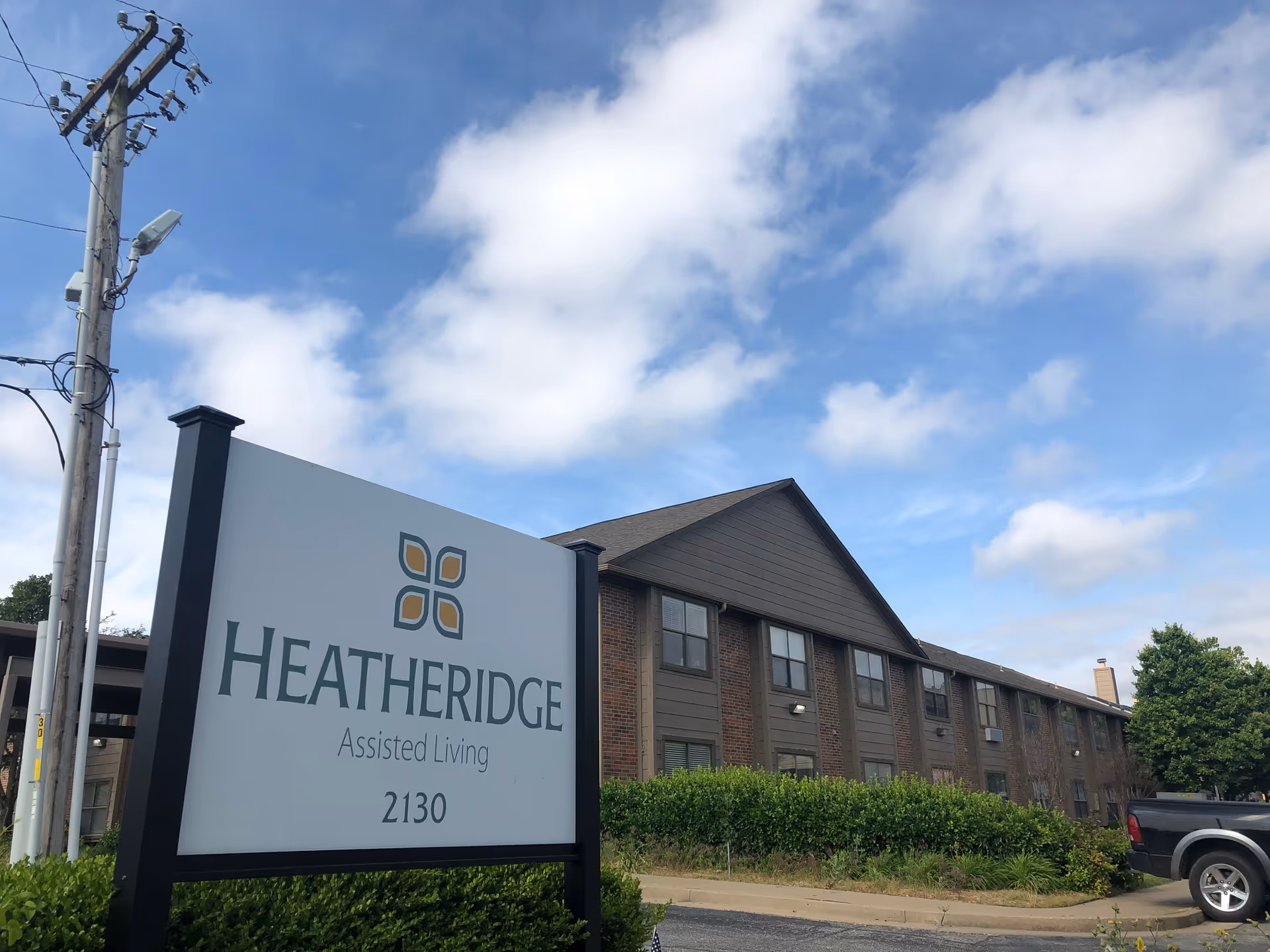 Exterior view of the Heatheridge Assisted Living building with a large entrance sign reading 'Heatheridge Assisted Living 2130' under a partly cloudy sky.