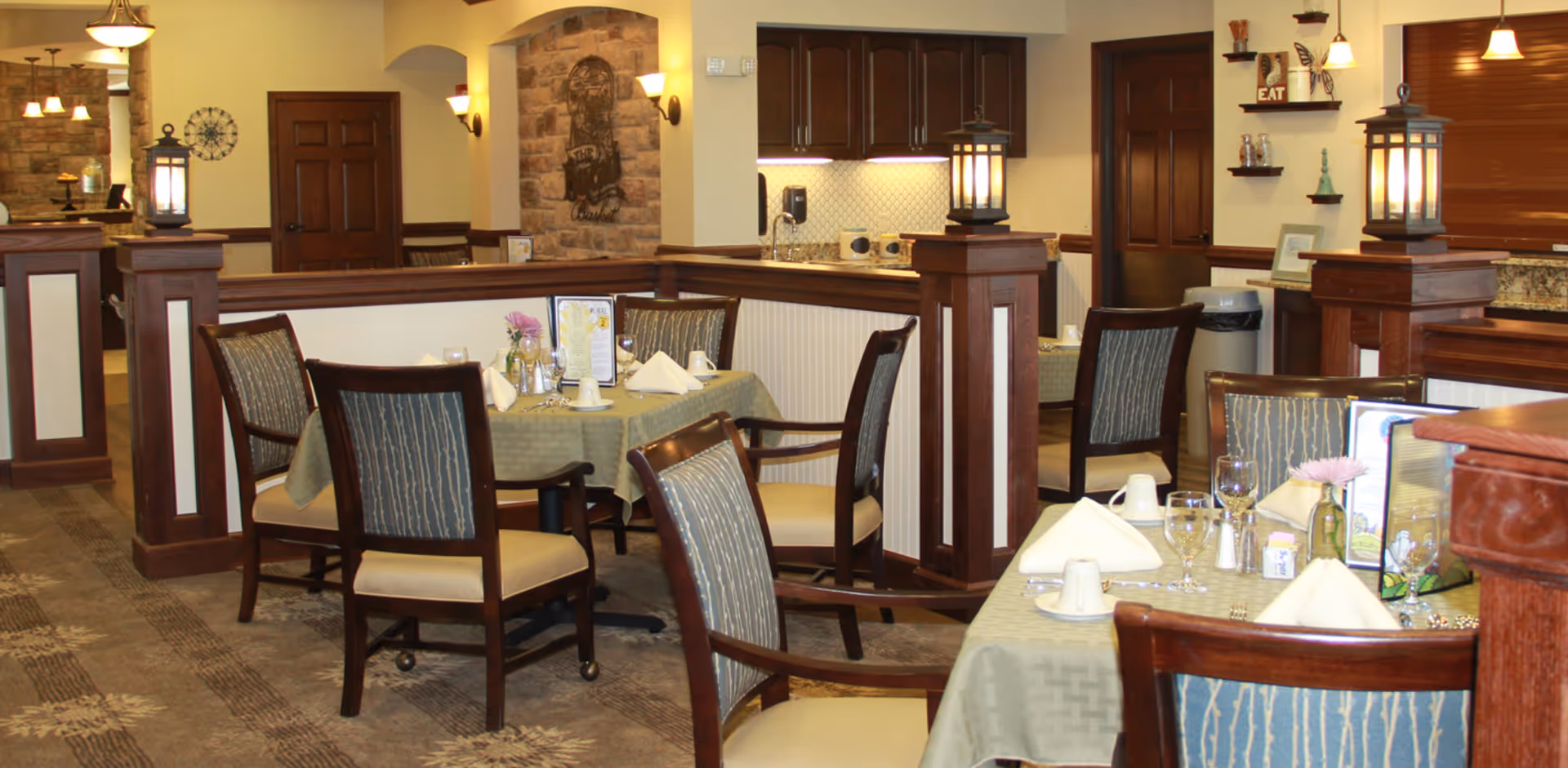 A dining area in a senior living facility with tables covered in green tablecloths, set with napkins, glasses, and cups. The room features wooden chairs with cushioned seats, decorative wooden pillars with lantern-style lights, and a warm, inviting interior with soft lighting and a stone accent wall in the background.