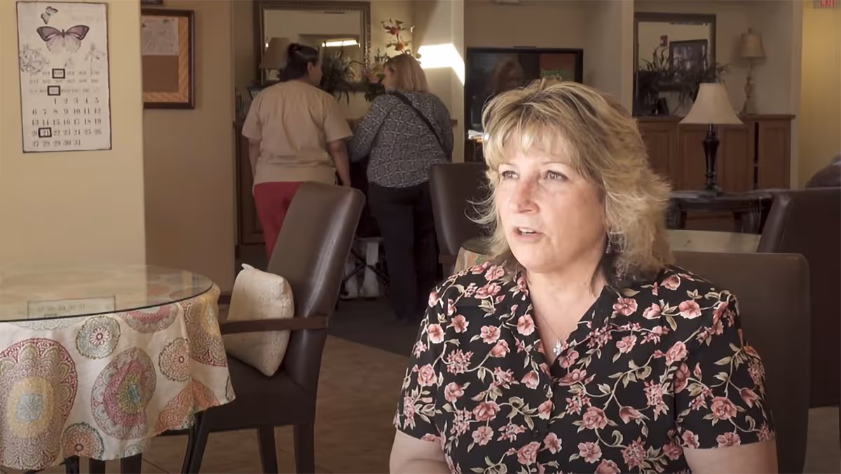 A woman with blonde hair wearing a black floral blouse is seated in a room with brown chairs and a round table covered with a patterned tablecloth. In the background, two other women are walking away from the camera. The room has warm lighting and home-style decor including lamps and framed pictures.