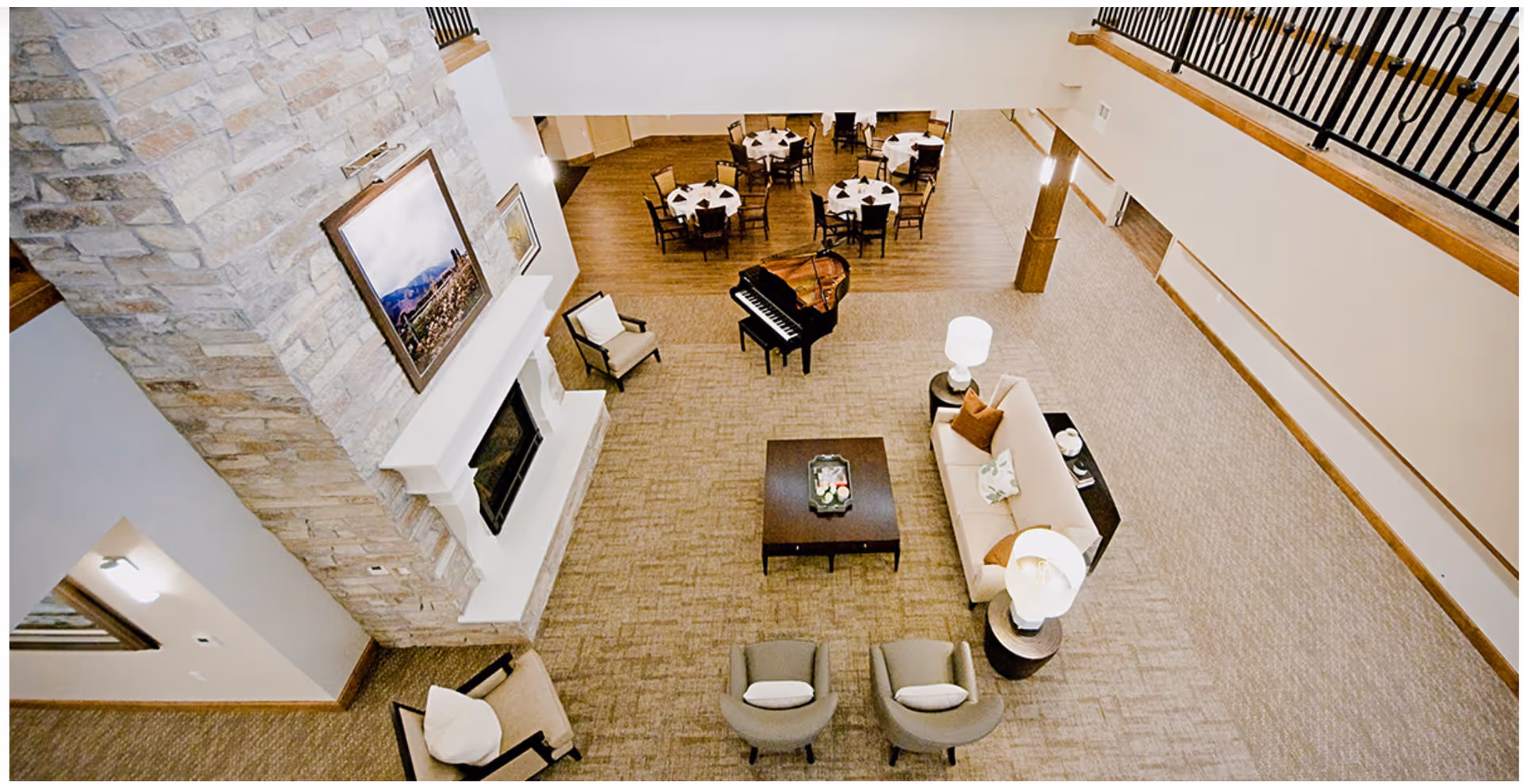 A spacious assisted living common area with a stone fireplace, a grand piano, several round dining tables with chairs, a beige sofa, armchairs, and lamps. The room has a high ceiling with a balcony railing visible above.