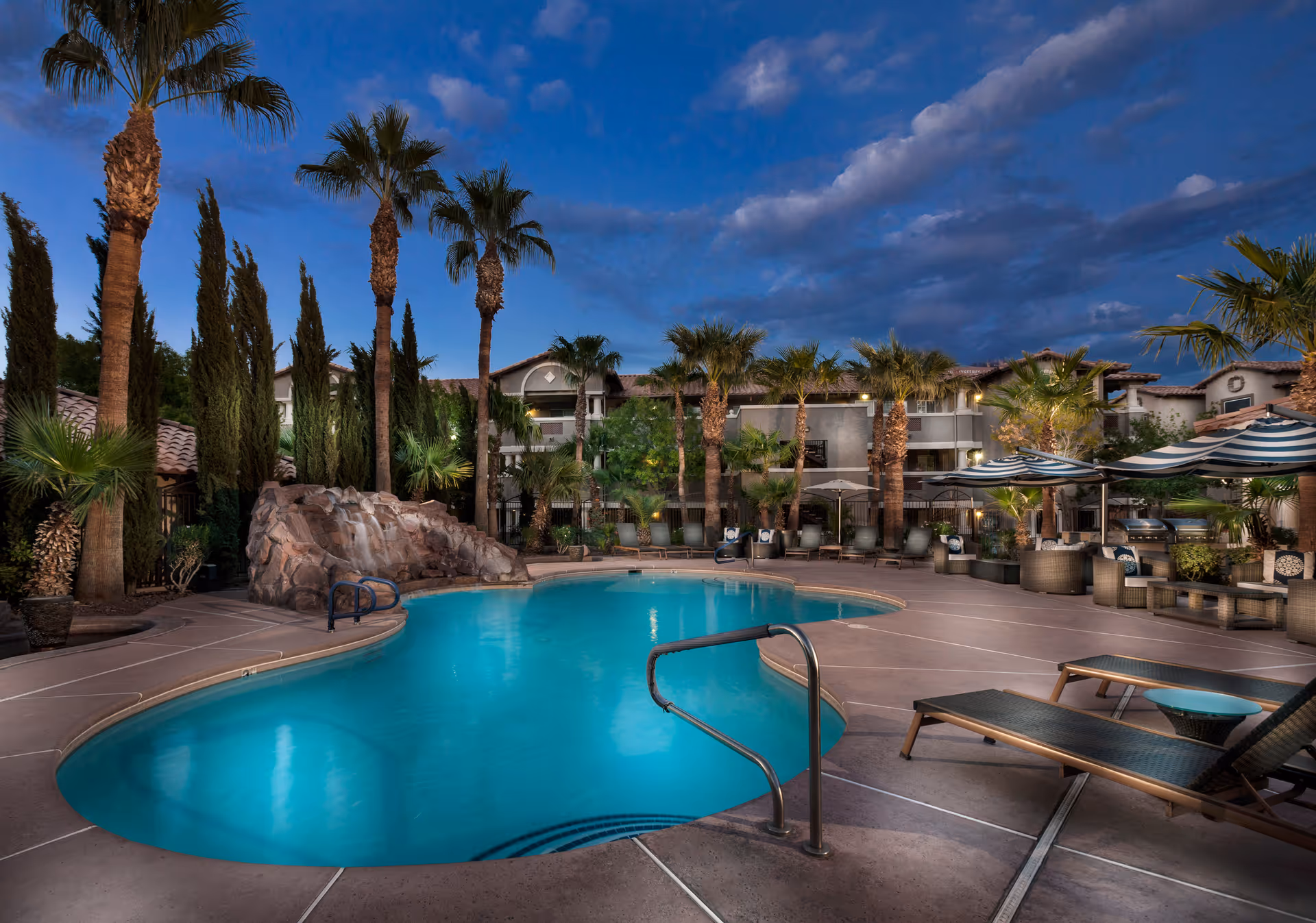 Outdoor swimming pool at dusk with palm trees and lounge chairs surrounding it. There is a rock waterfall feature on one side of the pool and striped umbrellas shading seating areas. A multi-story building is visible in the background under a partly cloudy sky.