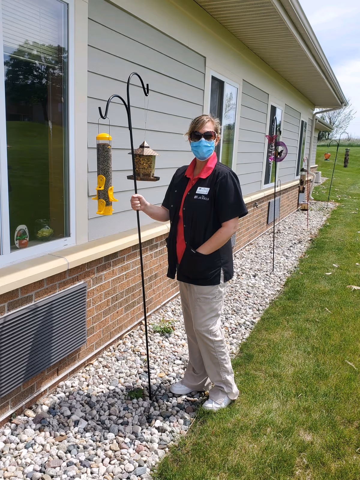 A woman wearing sunglasses, a face mask, a black jacket, red shirt, and beige pants is standing outside next to a building with gray siding and brick lower walls. She is holding a metal pole with bird feeders attached. The ground is covered with small rocks and there is green grass beside it.