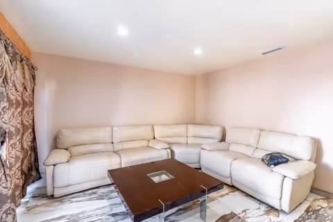 Bright living room with a large beige sectional sofa, a dark wooden coffee table, patterned curtains, and tiled floor.
