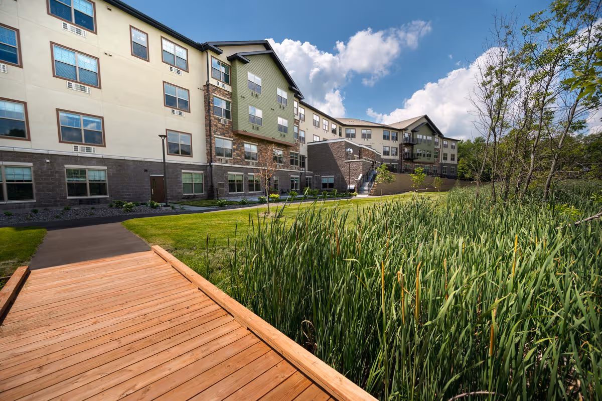 Exterior view of a multi-story senior living building with a wooden boardwalk, grassy lawn, and tall reeds under a blue sky.