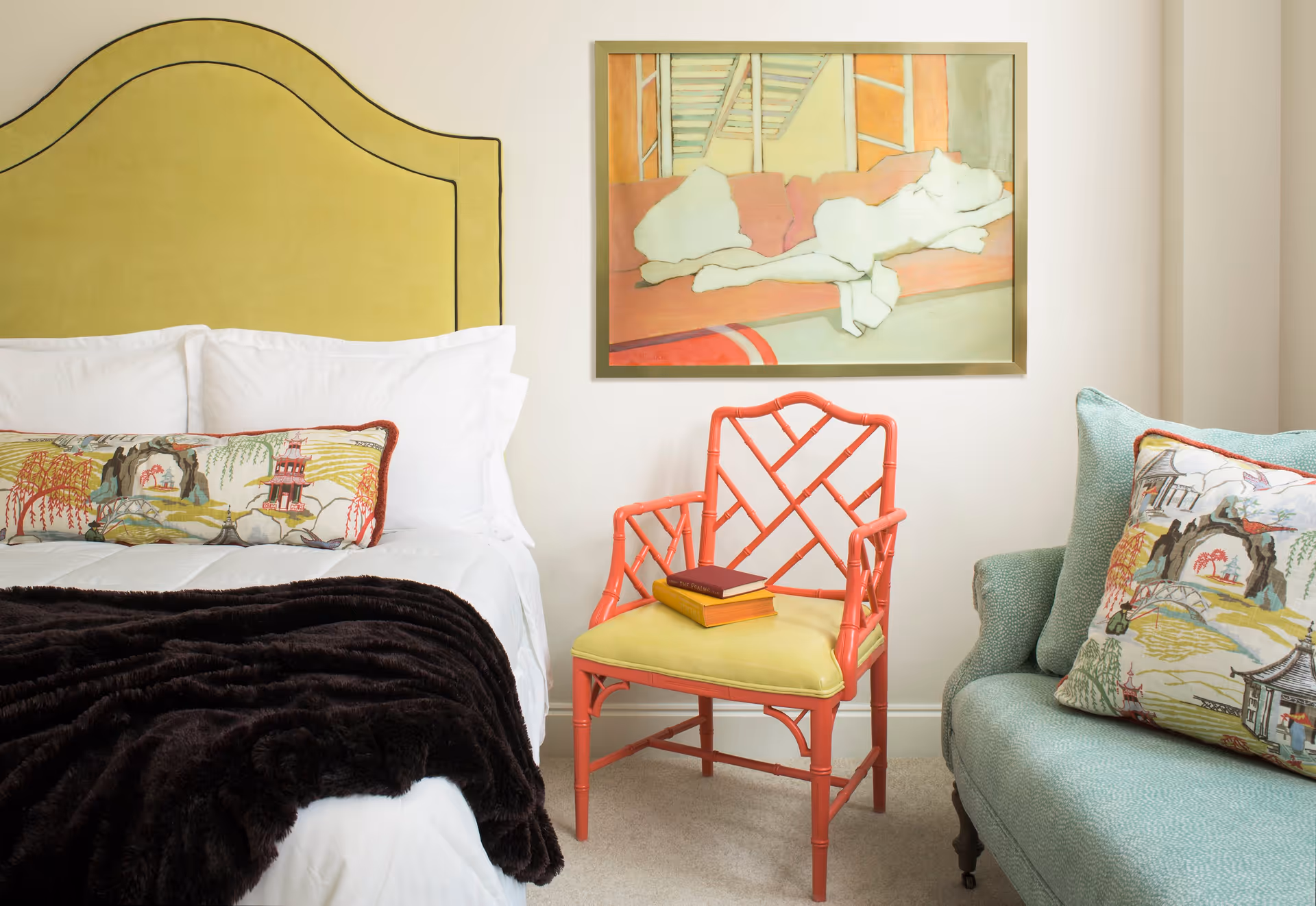 A cozy bedroom featuring a bed with white linens, a yellow upholstered headboard, and a decorative pillow with an Asian-inspired design. Next to the bed is a coral-colored chair with a yellow cushion and two books on it. A light blue sofa with a matching decorative pillow is partially visible. Above the chair hangs a framed abstract painting with soft pastel colors.