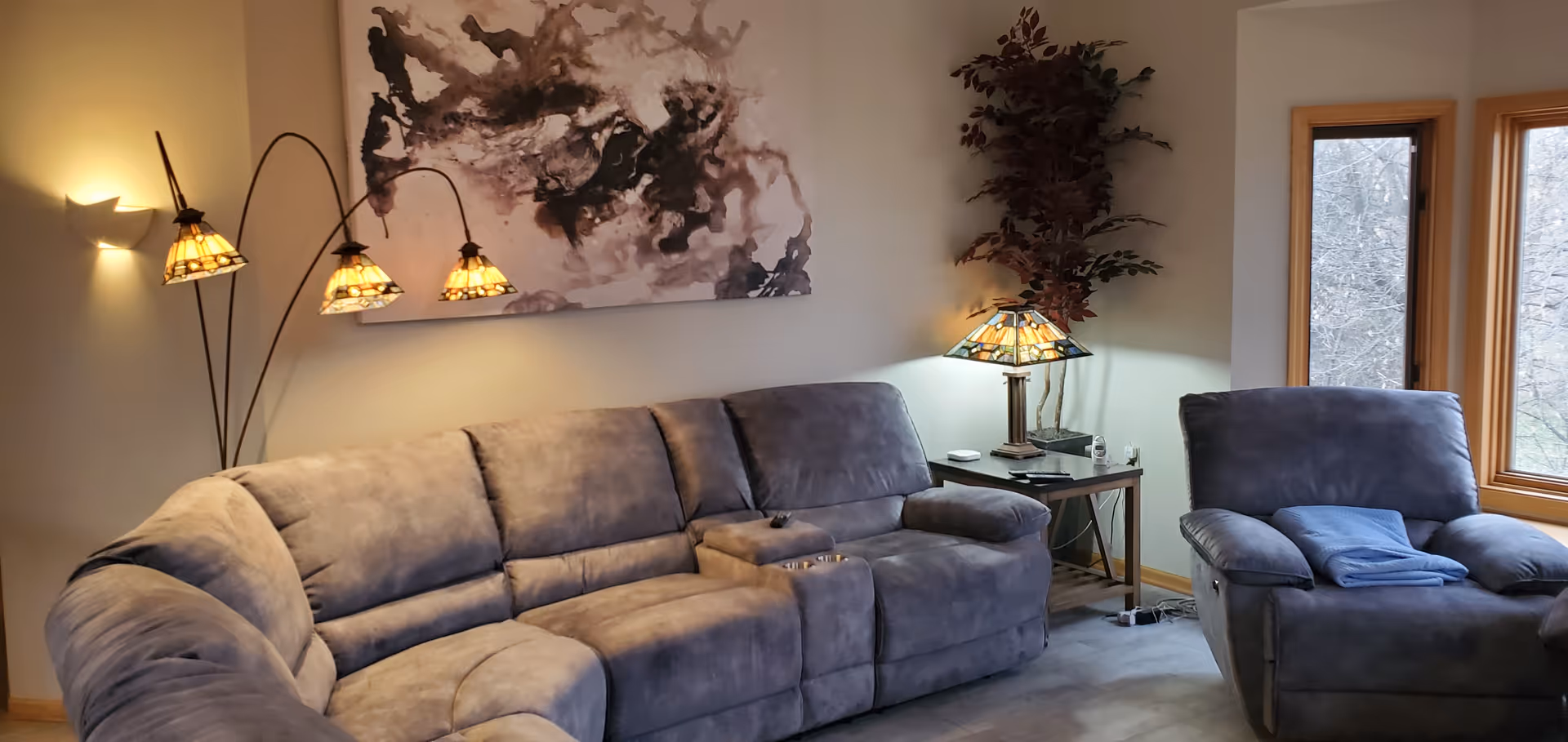 A cozy living room with a large gray sectional sofa and a matching armchair. There is a side table with a decorative lamp and a potted plant in the corner. A modern floor lamp with three stained glass shades is positioned behind the sofa. Two windows with wooden frames let in natural light, and an abstract painting hangs on the wall above the sofa.