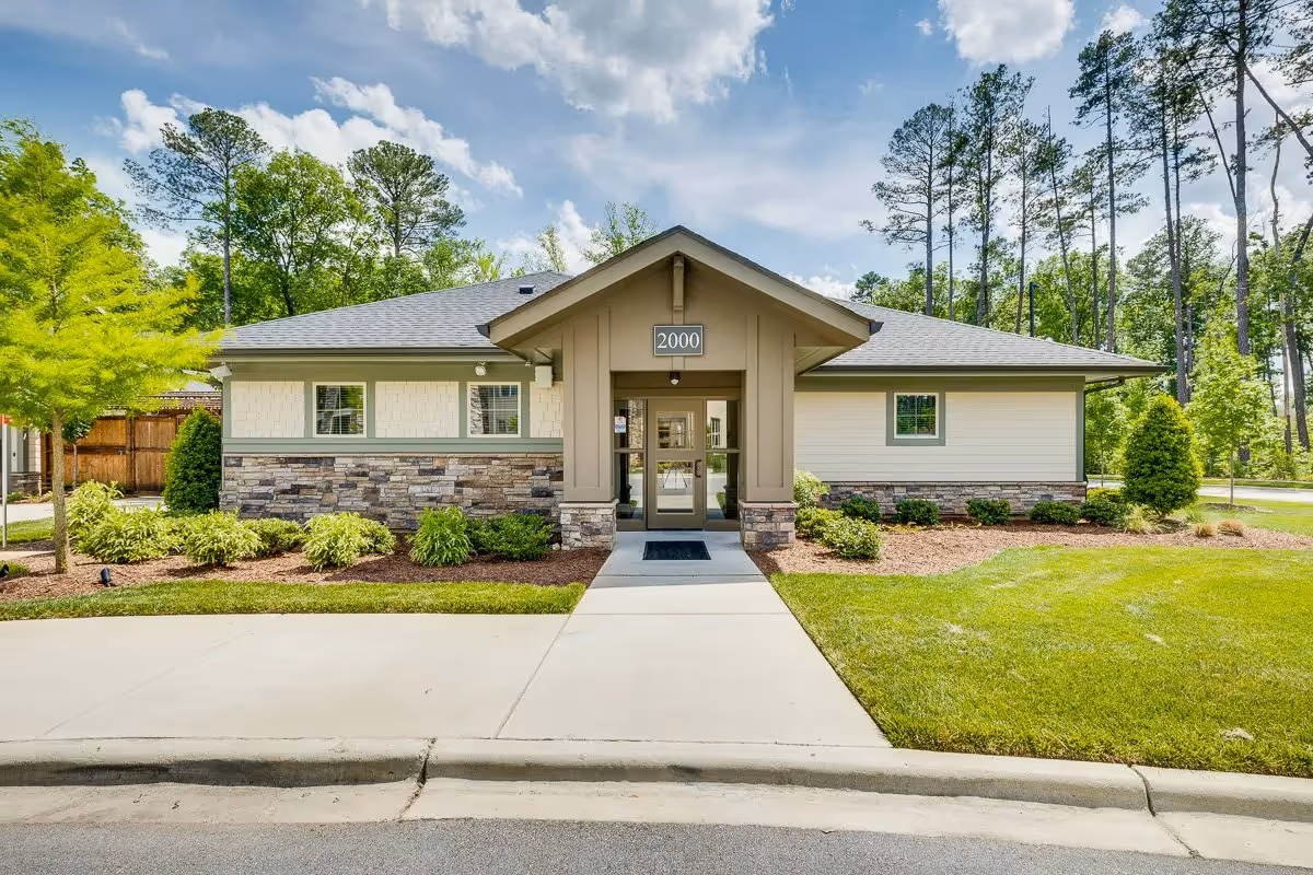 Single-story community building with a central glass entrance labeled "2000", stone and siding exterior, and landscaped lawn and trees under a partly cloudy sky.