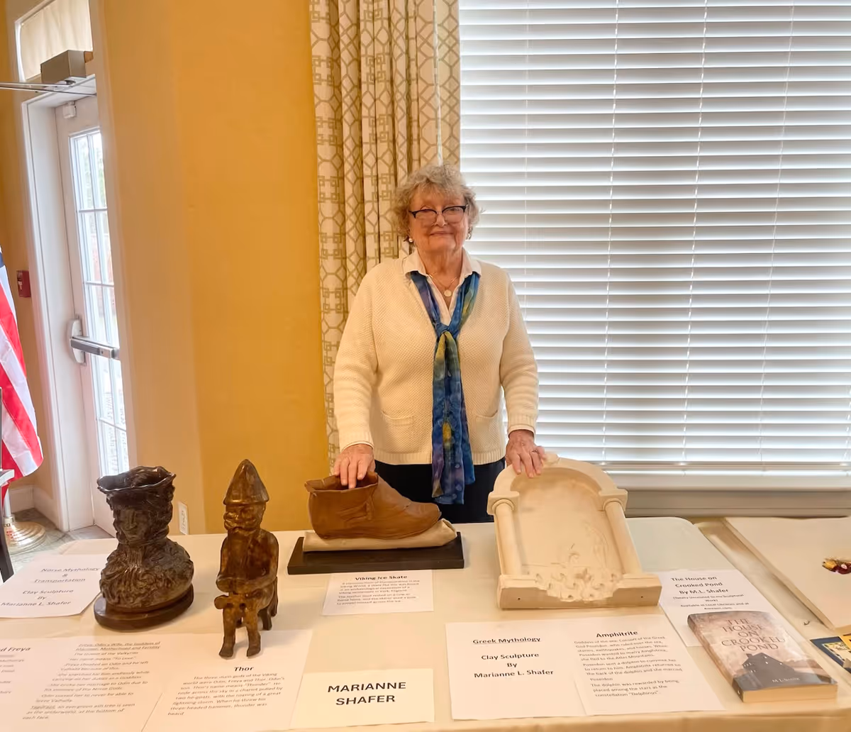 An elderly woman with glasses and a white sweater stands behind a table displaying various clay sculptures and informational placards in a well-lit room with a window covered by blinds and patterned curtains.