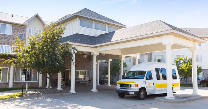 Front entrance of a senior living facility with a covered porte-cochère and a white shuttle van parked underneath.