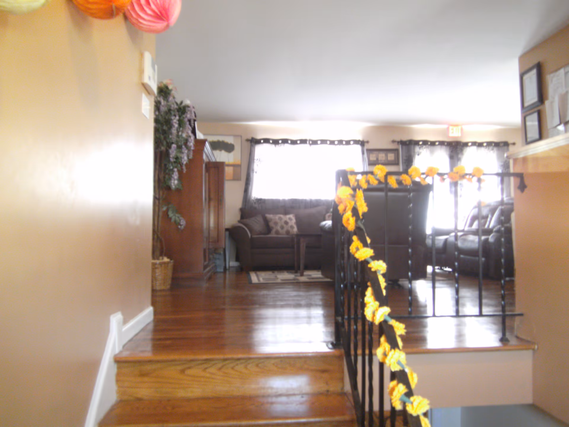 Interior view of a living room area with wooden flooring and a few steps leading up to it. The room has brown sofas, a wooden cabinet, and windows with dark curtains letting in bright light. A black metal railing decorated with yellow flowers lines the steps. The walls are painted beige, and there are framed certificates or documents on the wall to the right.