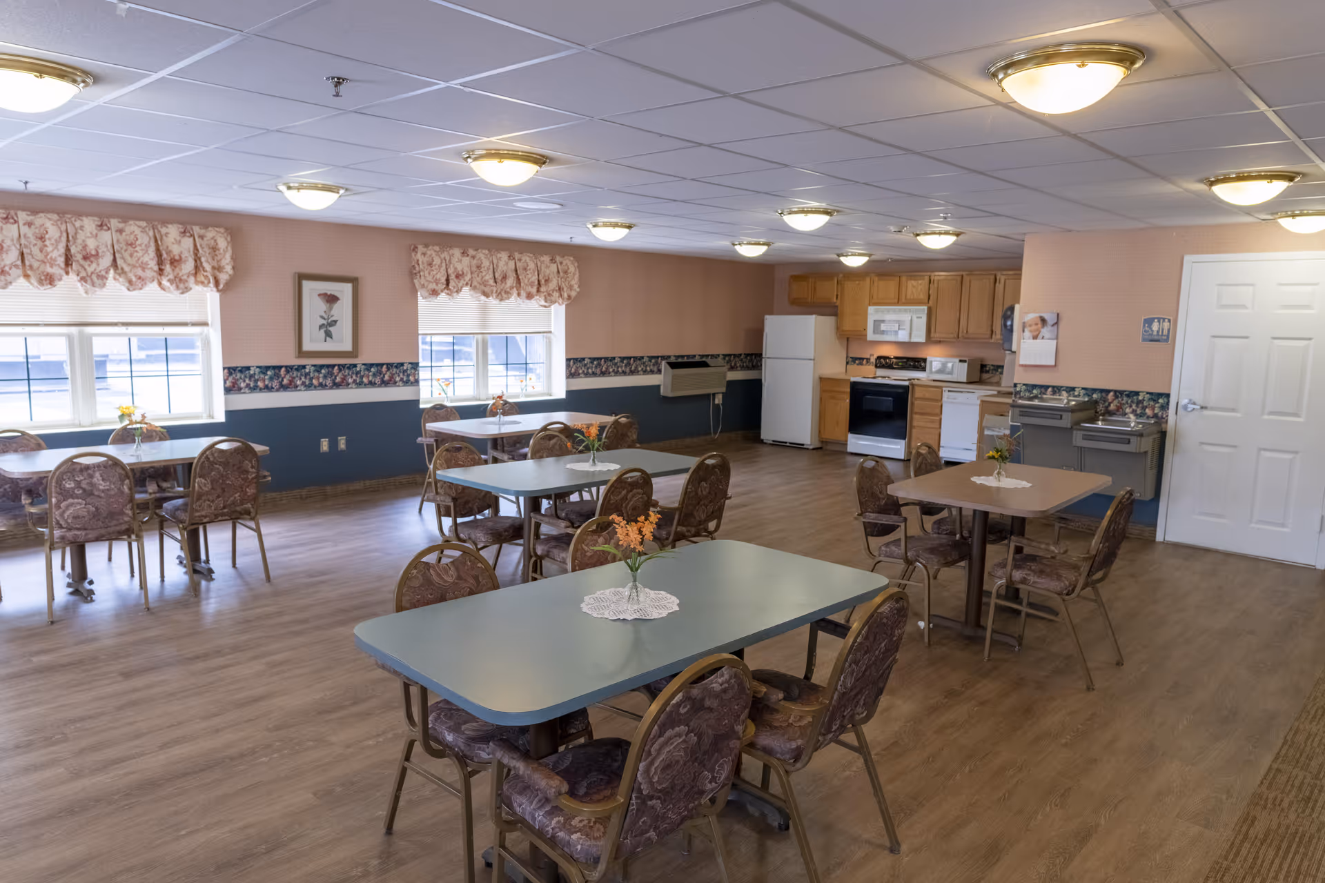 A dining room in a senior living facility with several tables and chairs arranged neatly. Each table has a small vase with orange flowers and a doily. The room has wood flooring, floral wallpaper border, and windows with floral valances. In the background, there is a kitchen area with a refrigerator, stove, microwave, and cabinets. Water fountains and a restroom sign are visible near a white door.