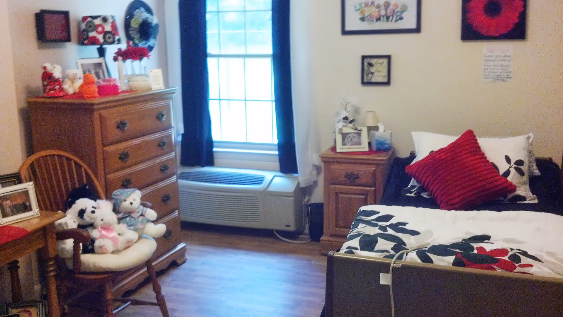 A cozy bedroom in a senior care community featuring a single bed with floral bedding and a red pillow, a wooden nightstand with a lamp and framed photos, a wooden dresser with decorative items on top, a wooden chair with stuffed animals, and a window with dark curtains and an air conditioning unit below it.