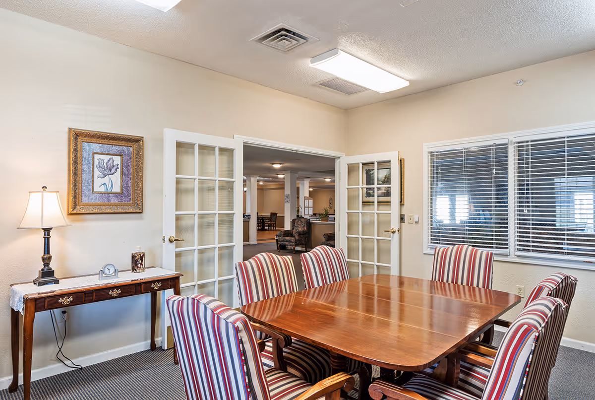 A meeting or dining room in a senior living facility with a wooden table surrounded by six striped upholstered chairs. There is a wooden side table with a lamp, a clock, and a decorative item against the wall. French doors open to a larger common area with additional seating and tables. A window with blinds is on the right wall.