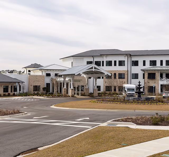 Exterior view of Watercrest Myrtle Beach senior living facility showing a large building with multiple windows, a covered entrance with white pillars, a roundabout driveway, landscaped areas with grass and shrubs, and a black fountain near the entrance.