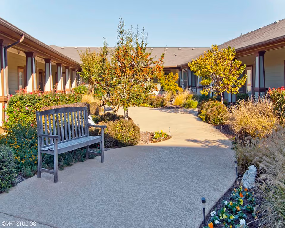 Courtyard walkway with a wooden bench, landscaped plants and trees surrounded by single-story assisted living buildings.