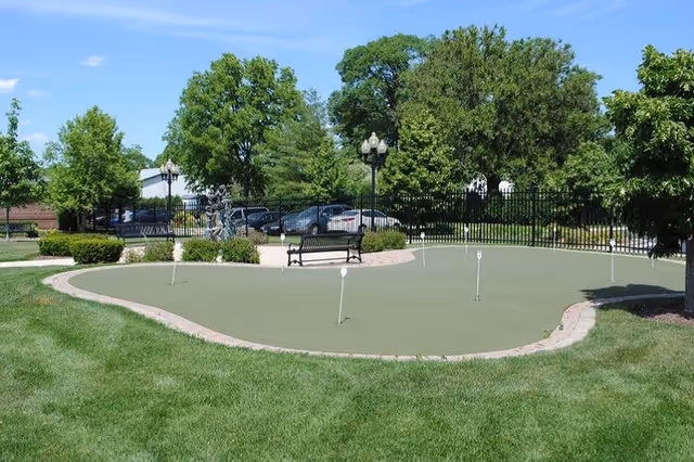 Outdoor putting green area with several golf holes, surrounded by well-maintained grass, trees, and bushes. There is a black bench and a decorative lamp post near the putting green, with a parking lot and cars visible in the background under a clear blue sky.