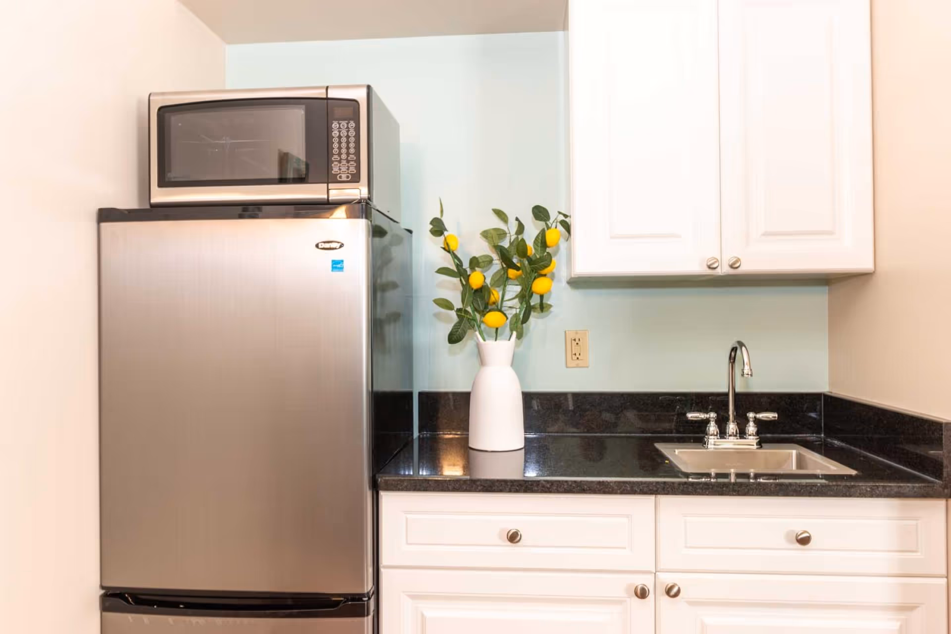 Compact kitchenette with a stainless-steel refrigerator topped by a microwave, white cabinets, black countertop with a sink, and a vase of lemons.
