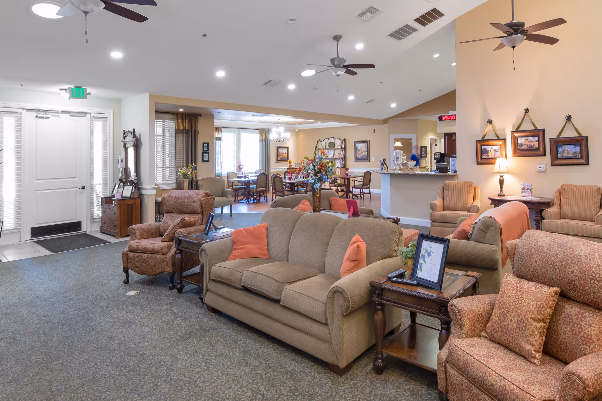A spacious and well-lit assisted living facility common area with comfortable seating including sofas and armchairs arranged around side tables. The room features ceiling fans, framed artwork on the walls, and a dining area with tables and chairs in the background. There is a reception or service counter visible, and the entrance door is on the left side of the image.